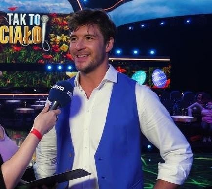 Mikołaj Roznerski w swobodnym stroju, zamyślony, na tle wiejskiego krajobrazu, w otoczeniu zieleni.