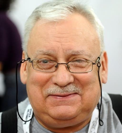 Szczegółowe ujęcie książek Andrzeja Sapkowskiego, w tym cyklu o Wiedźminie i trylogii husyckiej, ułożonych na półce lub biurku.