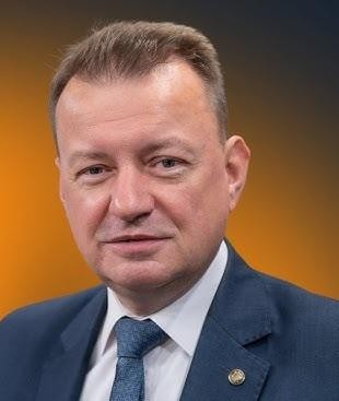 Mariusz Błaszczak podczas konferencji prasowej, przemawiający przy mównicy, w tle subtelne elementy związane z Ministerstwem Obrony Narodowej.