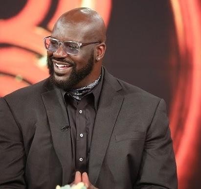 Shaquille O'Neal w garniturze z mikrofonem na scenie, uśmiechnięty.