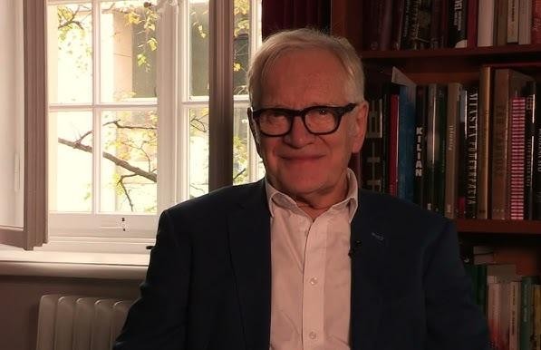 Andrzej Seweryn na scenie teatralnej, w kostiumie, z ekspresyjnym gestem, oświetlony światłami reflektorów.