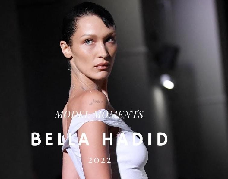 Bella Hadid za kulisami pokazu mody, w dynamicznej pozie, w otoczeniu przygotowań do wybiegu, ukazująca energię i glamur branży.